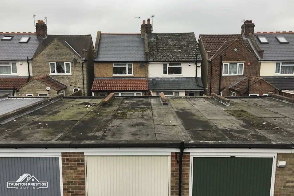 Row of flat-roofed brick lock-up garages with worn felt below semi-detached houses